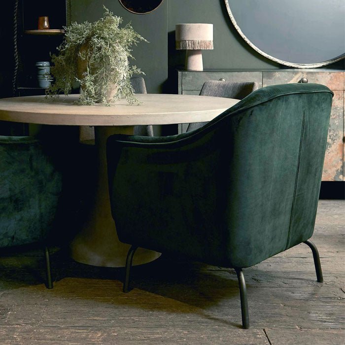 The curved back of a green velvet armchair next to a dining table.