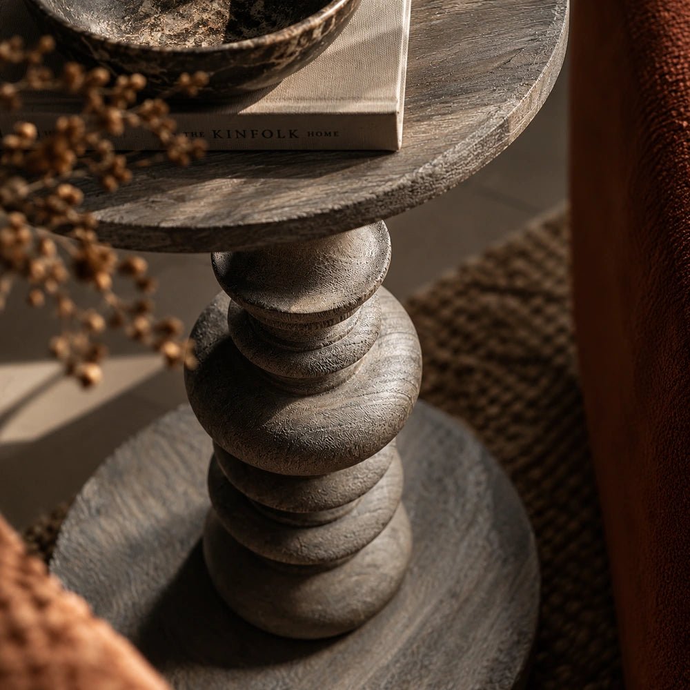 Close-up of the Devon Wooden Side Table showing its sculptural stacked pedestal, rounded tabletop edge and dark textured finish within a warmly styled interior.