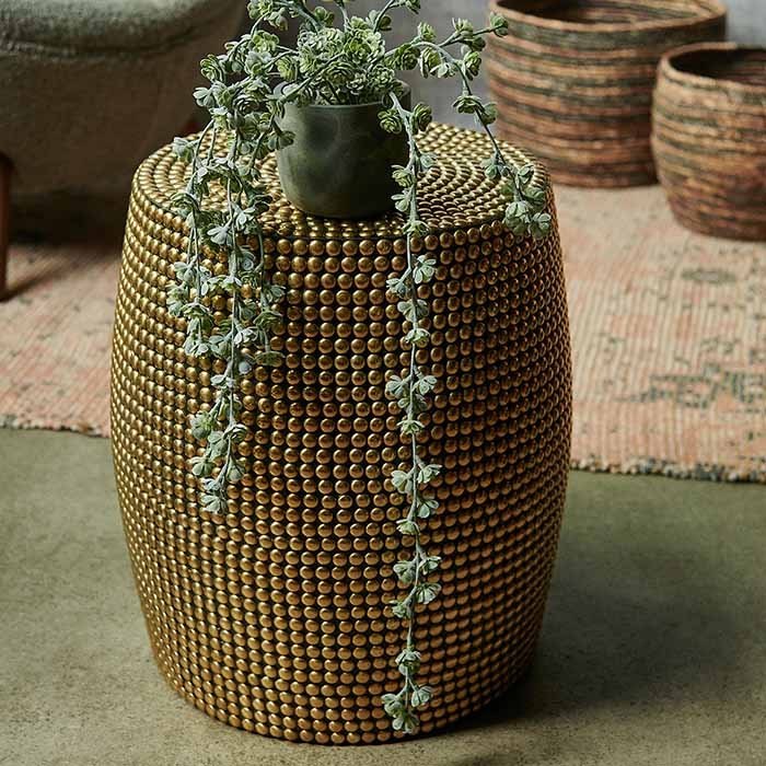 The Dewitt Brass Stud Stool, a tall round stool covered entirely in small brass studs, styled with a faux trailing plant.