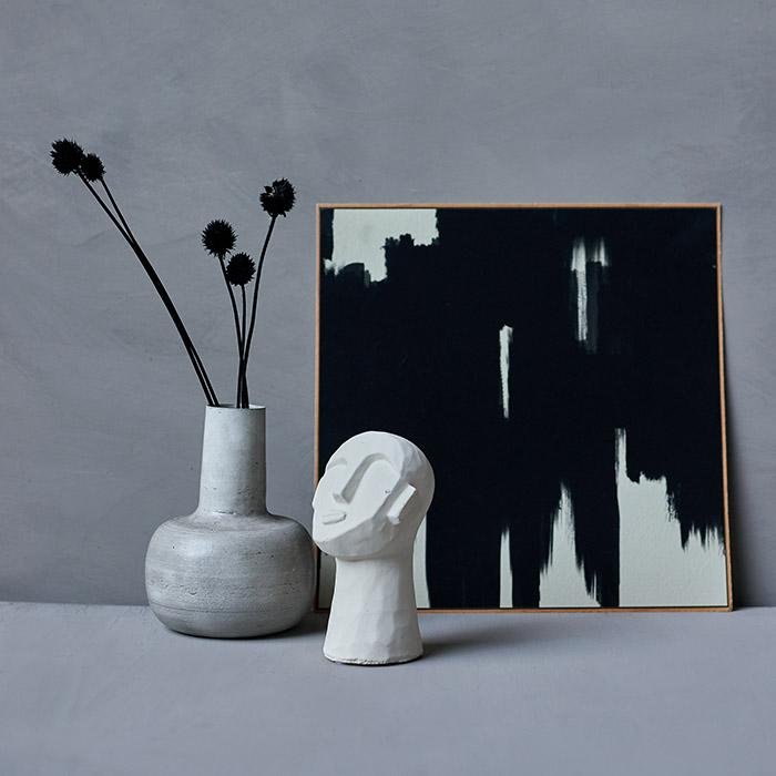 Square framed monochrome print leaning on a wall behind a face sculpture and vase