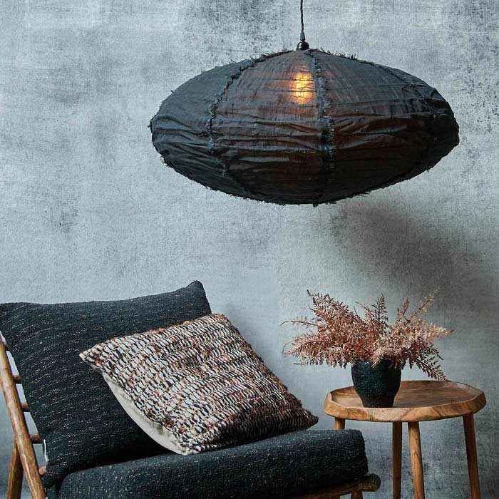 Large oval shaped black fabric pendant light switched on above a wooden side table and armchair. Luxury lighting to add designer style to your home. 