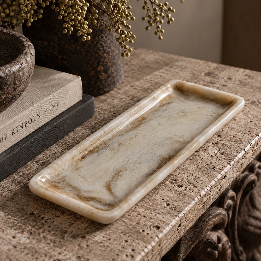Emery Rectangular Tray shown close up with pale marbled surface and raised rim, styled on a textured console beside stacked books and decorative vessels.