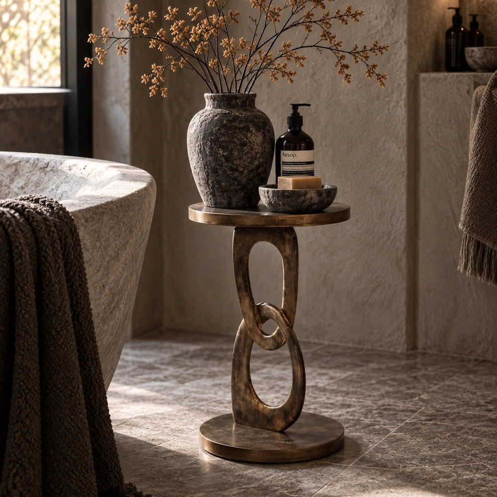 Emilio Side Table with sculptural linked pedestal styled beside a bed, topped with a textured vase, bottles and small decorative objects in a warm neutral bedroom.