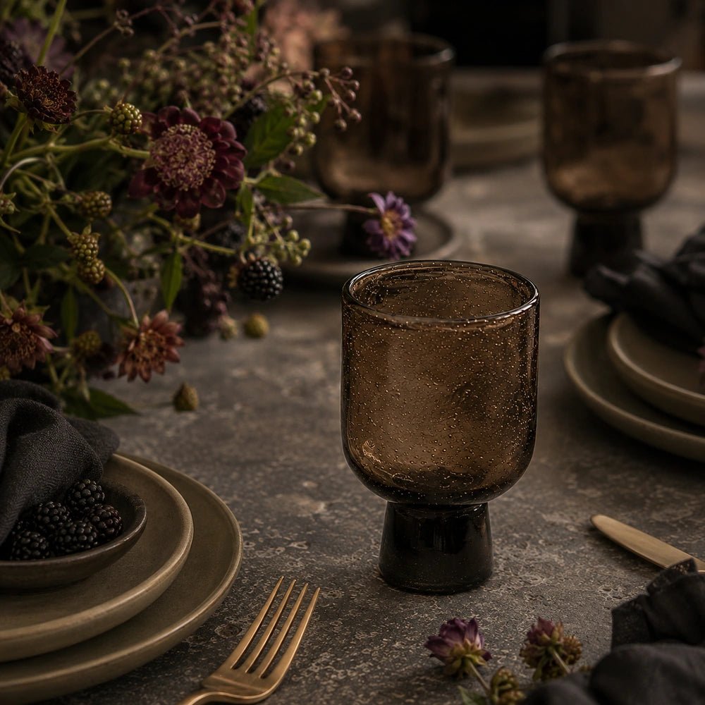 Evie Glass, Burnt Umber arranged in a layered dining table setting with plates, dark cutlery, flowers and candlelight, highlighting its warm textured glass.