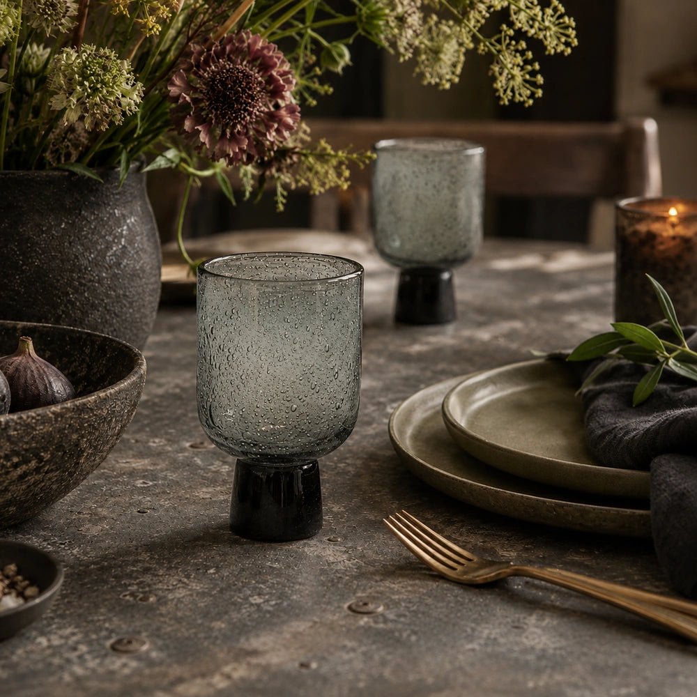 Evie Glass, Smoke with textured smoky glass bowl and dark pedestal base, styled among layered tableware, flowers and candlelight on a dining table.
