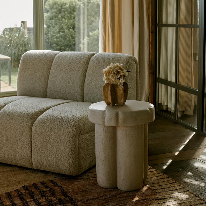 Styled image of a contoured and sculptural cream side table, with a small vase of fake flowers, and styled next to a large cream occasional chair.