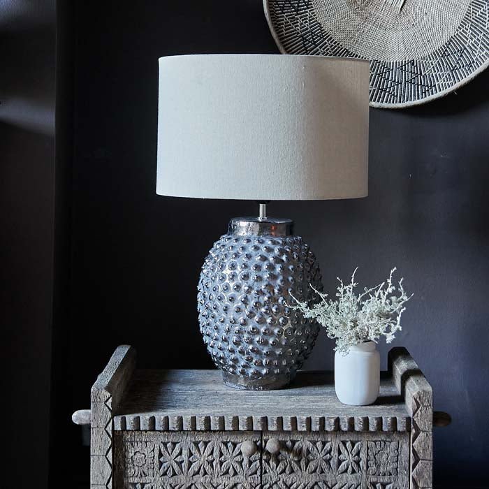 The Homer Table Lamp, a table lamp with a cream drum shade and metallic bobble-textured base, on a small console.