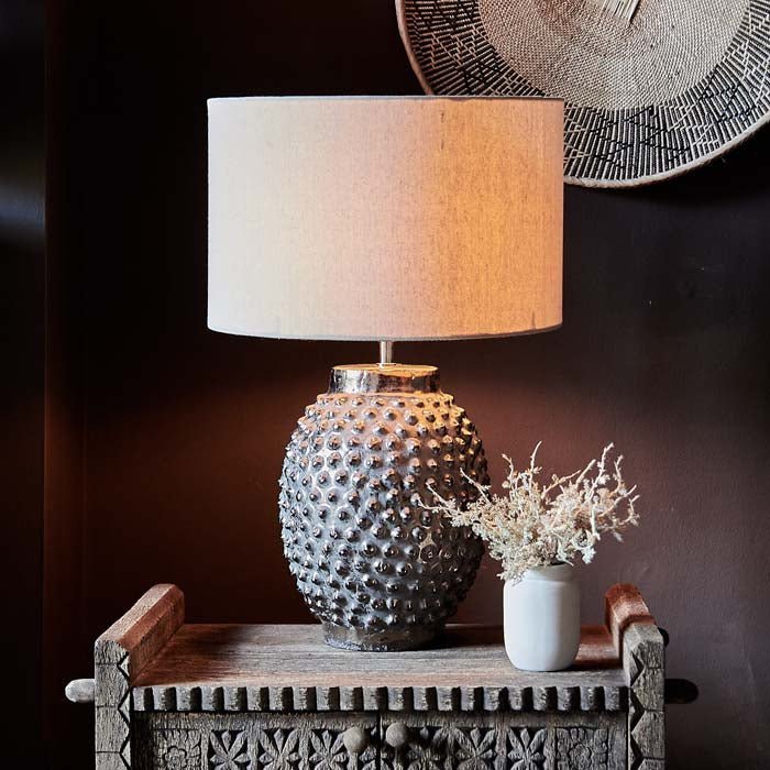 The Homer Table Lamp, a table lamp with a cream drum shade and metallic bobble-textured base, on a small console.