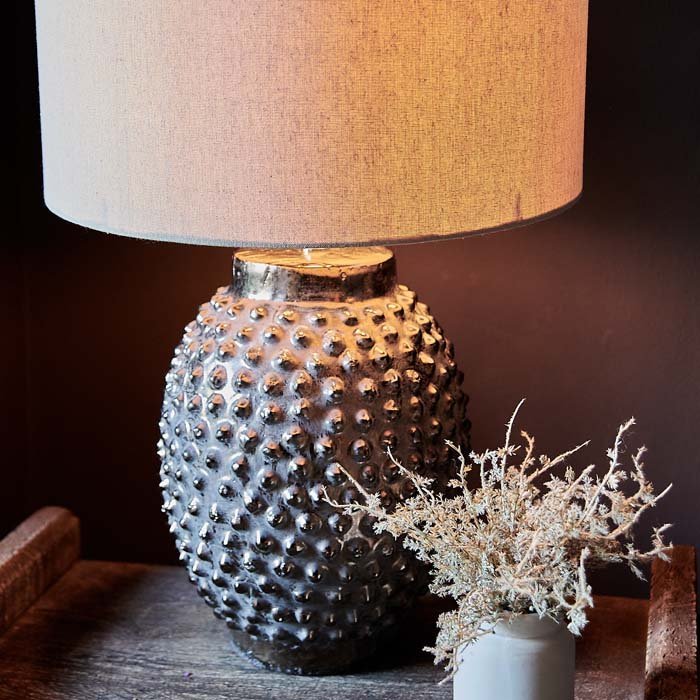 The Homer Table Lamp, a table lamp with a cream drum shade and metallic bobble-textured base, on a small console.
