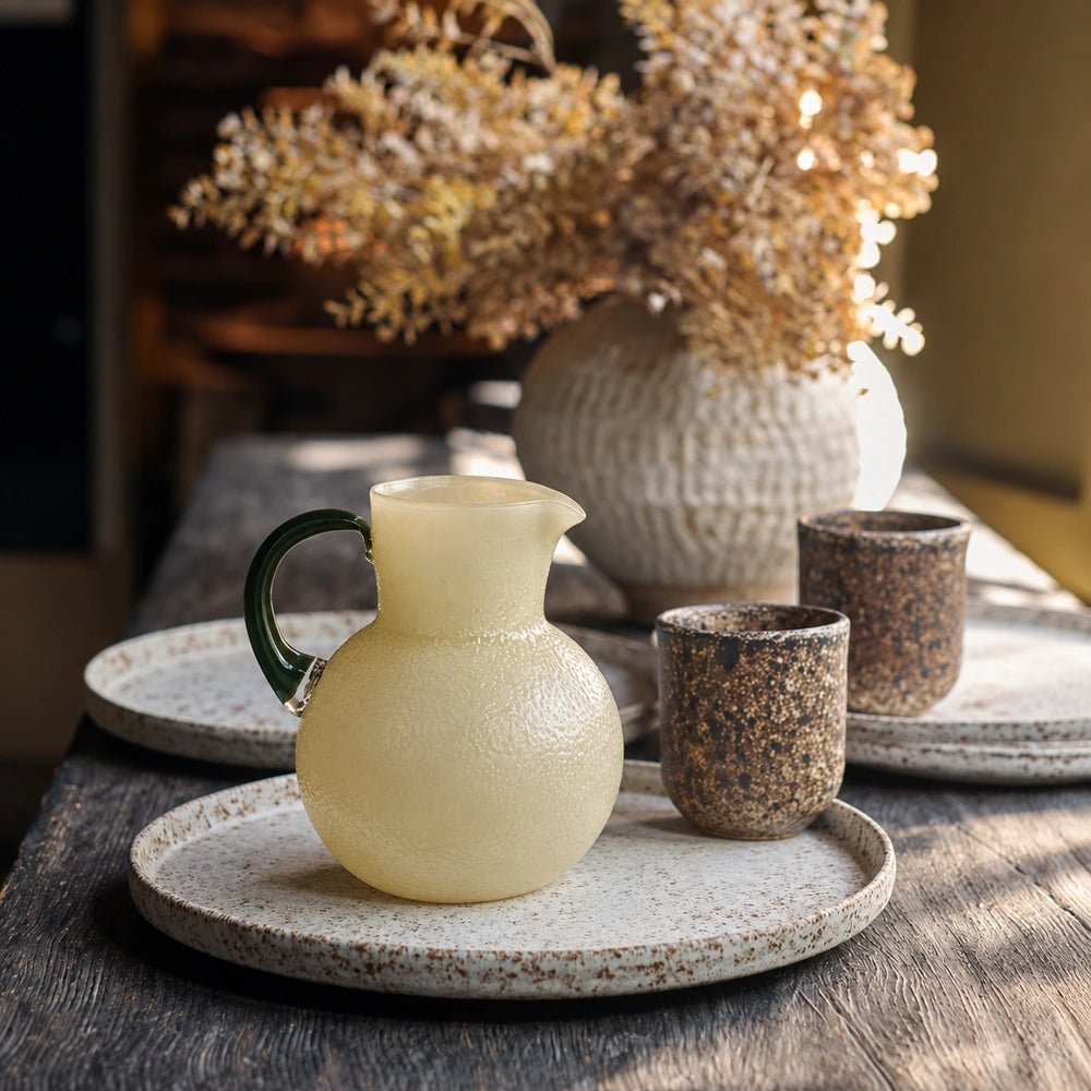 Limone Glass Jug with pale yellow textured glass and dark green handle, arranged with patterned cups, plates and dried flowers on a dining table.