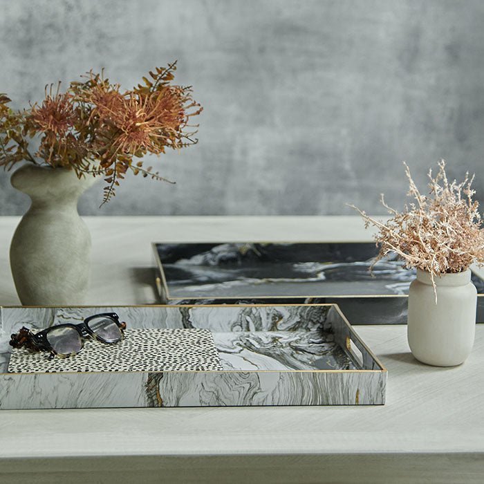 Magnus Trays - Set of 2, a set of two marble effect trays in two colorways black and white with brass details on a table.