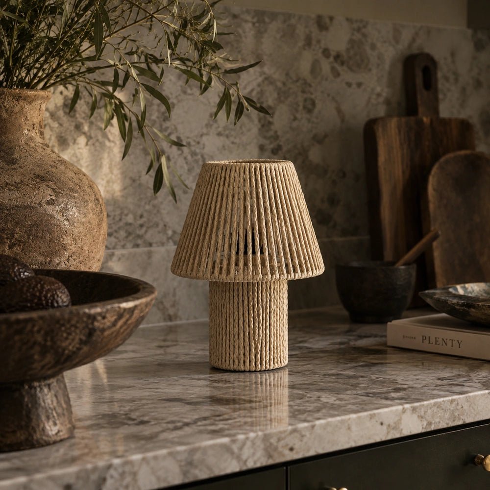 Malone Portable Table Lamp with textured woven string design, styled unlit on a marble kitchen counter among rustic vessels, foliage, wooden boards and bowls.