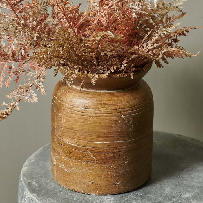Rustic textured brown ceramic vase sat on a round grey table with a faux bouquet of ferns