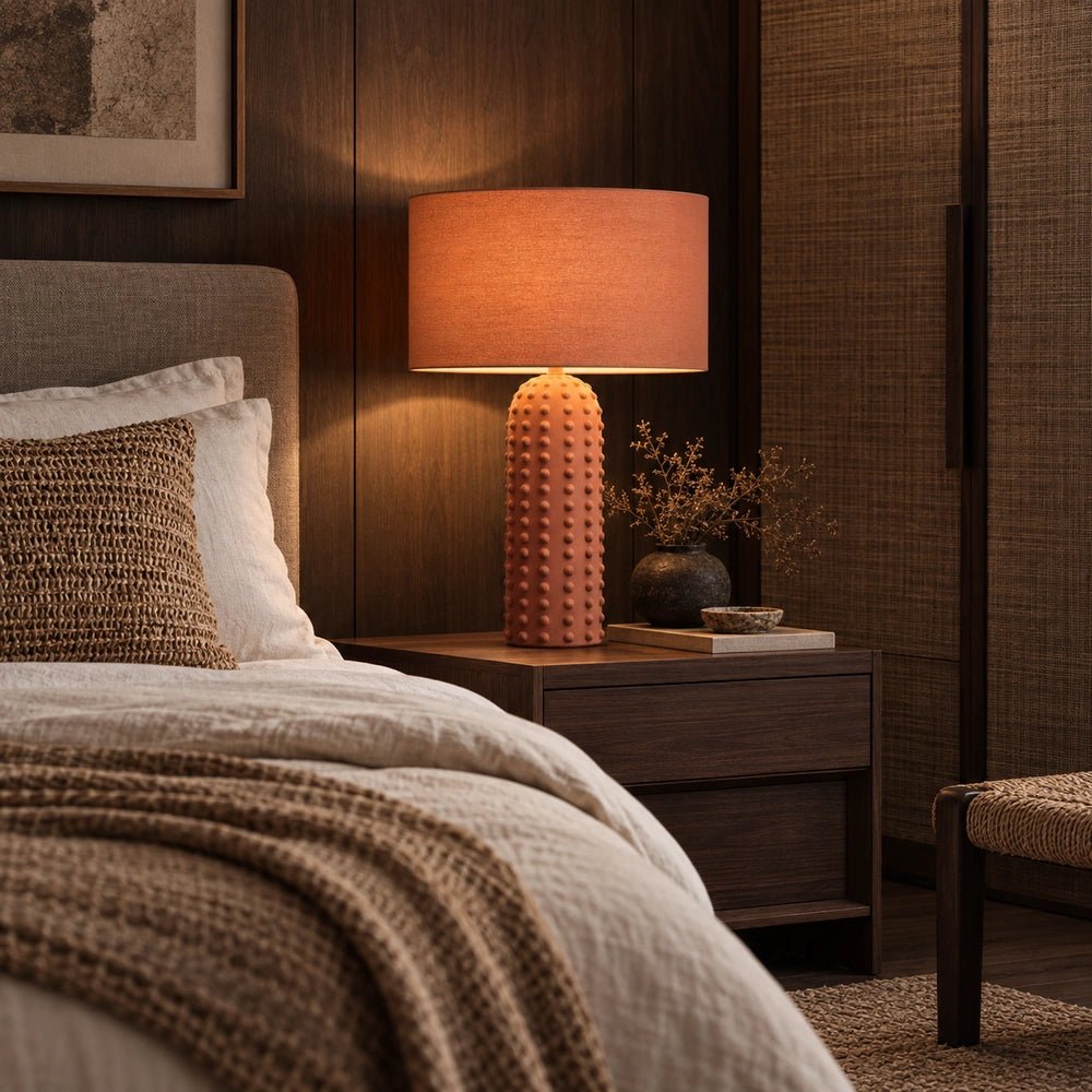 Maynard Terracotta Table Lamp with tall textured bobble base and wide fabric shade, illuminated on a wooden bedside table in a warm neutral bedroom.