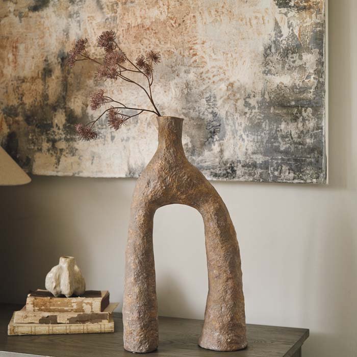 A sculptural, arched terracotta vase with two sides that join up at the opening, with a single artificial flower in it and sitting on a wooden console.