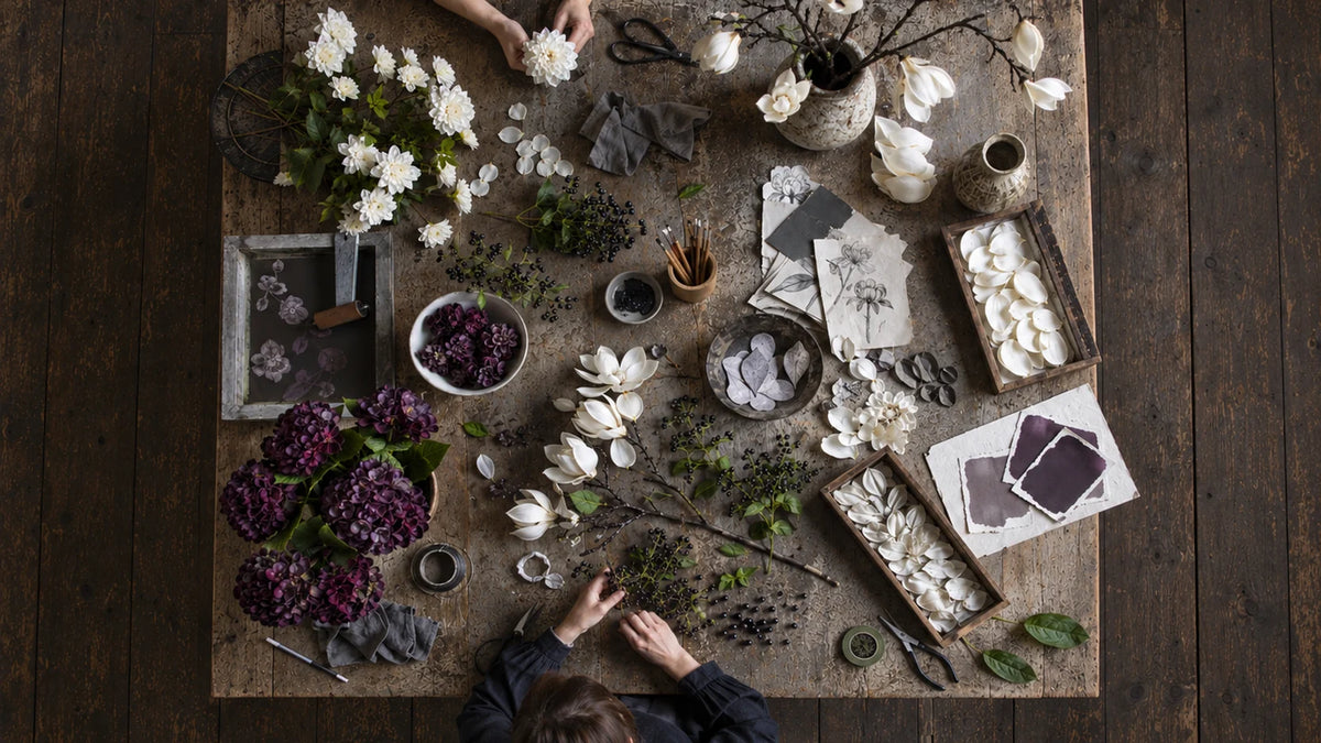 White magnolia branches and deep purple flowers arranged across a studio table with botanical sketches, petals, berries, vases, scissors and art materials.