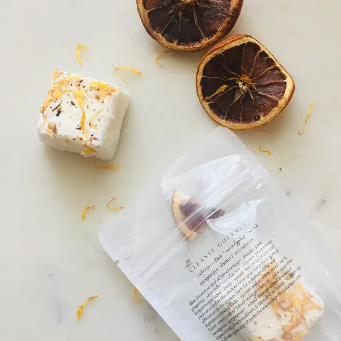 A square shower steamer topped with orange zest next to two dried slices and an open packet