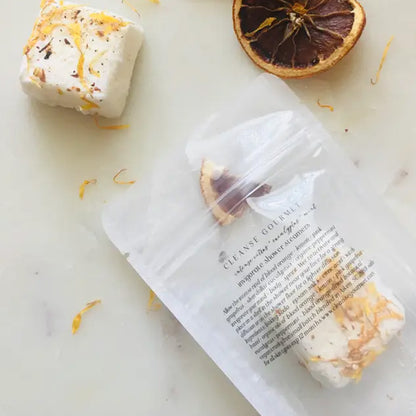 A white square shower steamer bar topped with orange zest next to a frosted plastic package with a steamer inside 