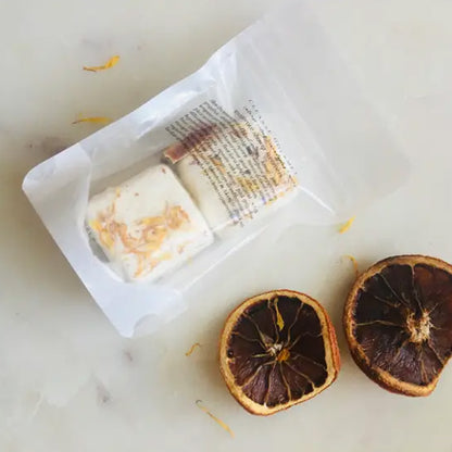 Two dried orange slices next to a closed plastic package containing two square shower steamer