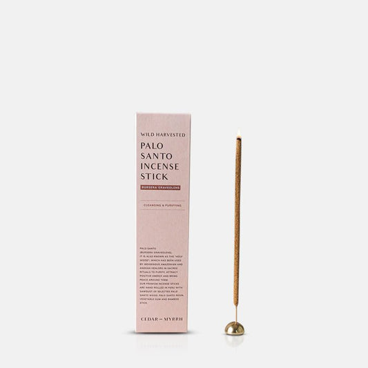 A single burning incense stick placed in a gold holder next to its pink packaging box
