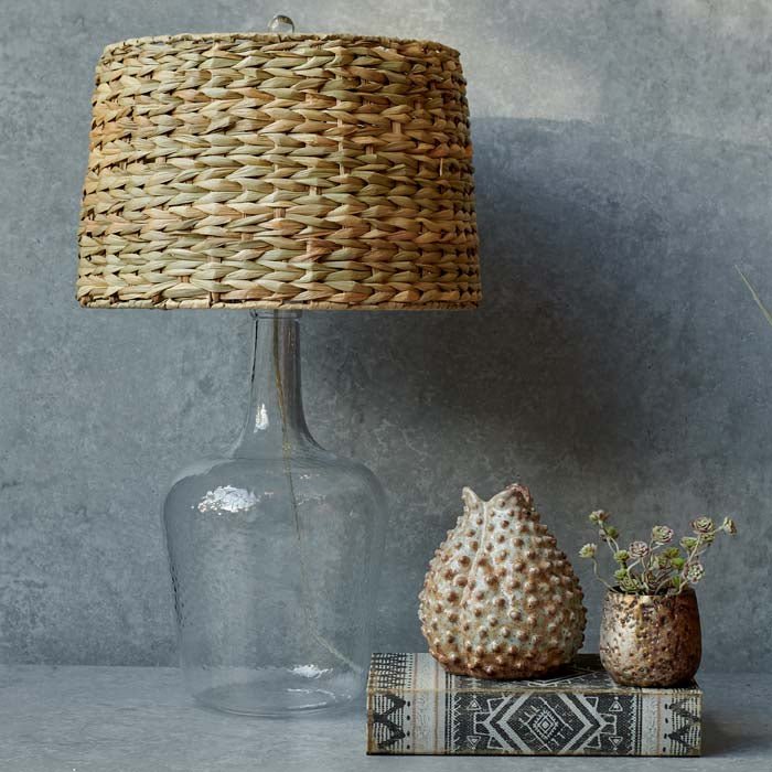 Clear glass base lamp with a round woven shade next to a small vase