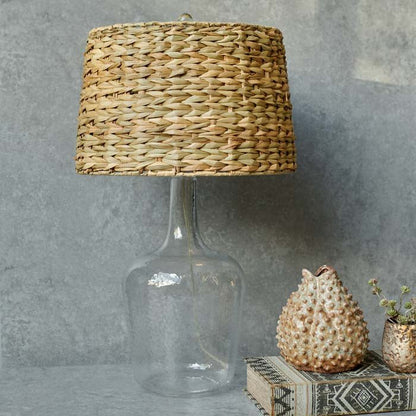 Tall clear glass table lamp with a bamboo shade sat next to a small textured vase