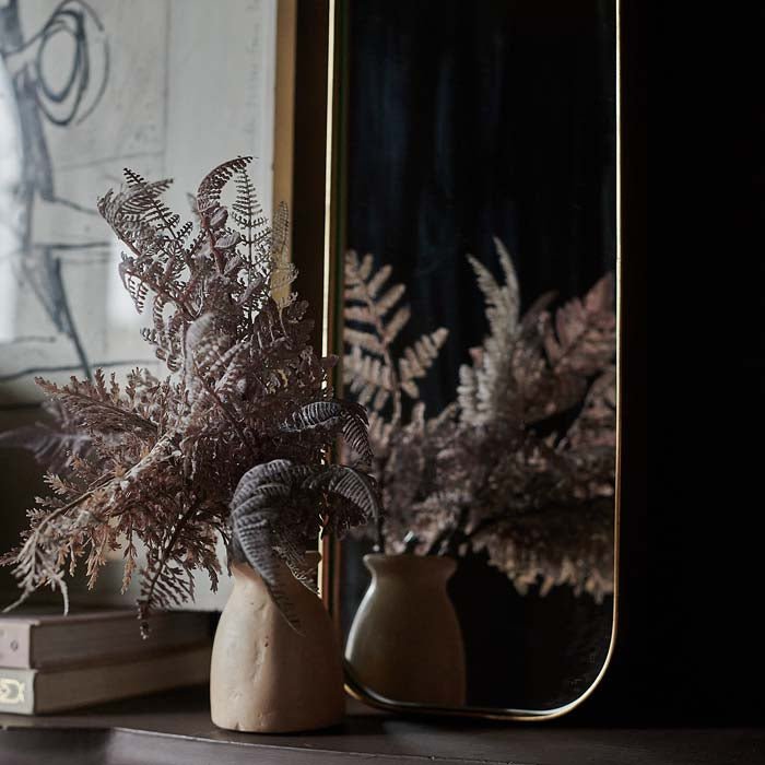 Curved gold frame around a rectangular mirror sat behind a bud vase with purple fern stems