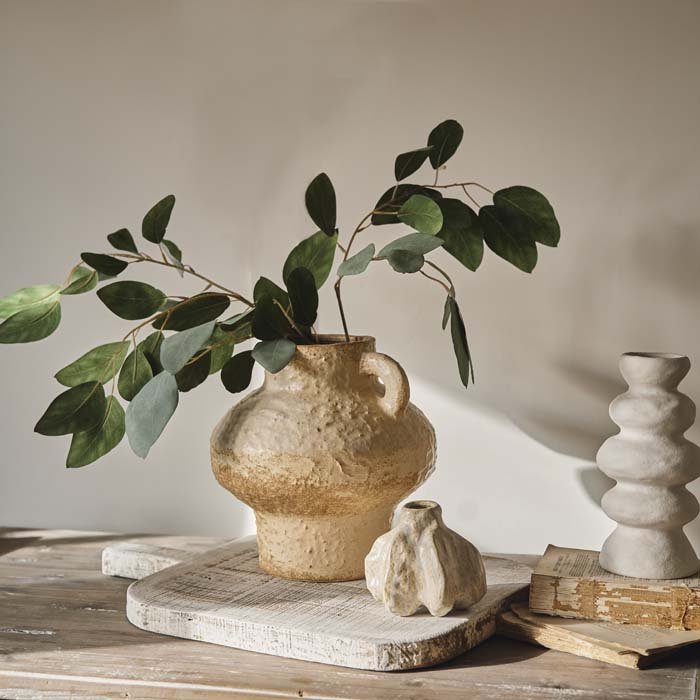 An artisan style vase with a cream ceramic textured glaze, styled with a stem of faux eucalyptus. Large organic style vase styled with a smaller artisan style bud vase