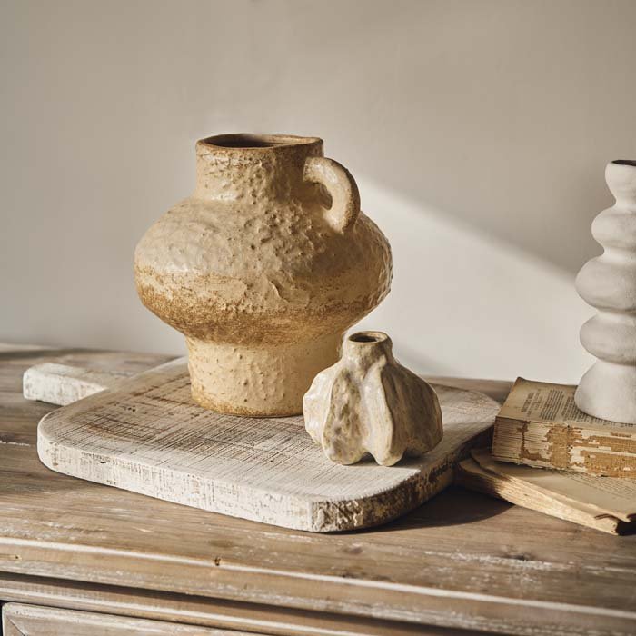 A cream shaped textured vase with a narrow neck and base and a wider middle and one handle. Reminiscent of a greek style vase with a cream bumpy texture styled with a smaller organic shaped bud vase. 