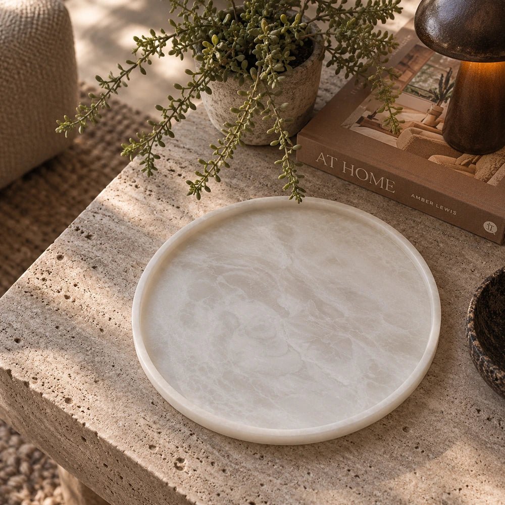 Salome Round Platter with pale marbled finish displayed on a textured coffee table beside books, foliage and warm neutral decorative accessories in a living room.