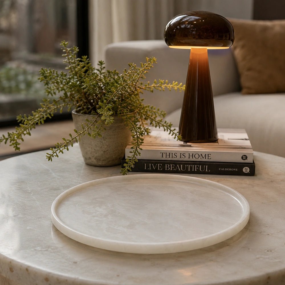 Salome Round Platter with pale marbled surface styled on a coffee table beside stacked books, a dark table lamp and leafy foliage in a neutral interior.