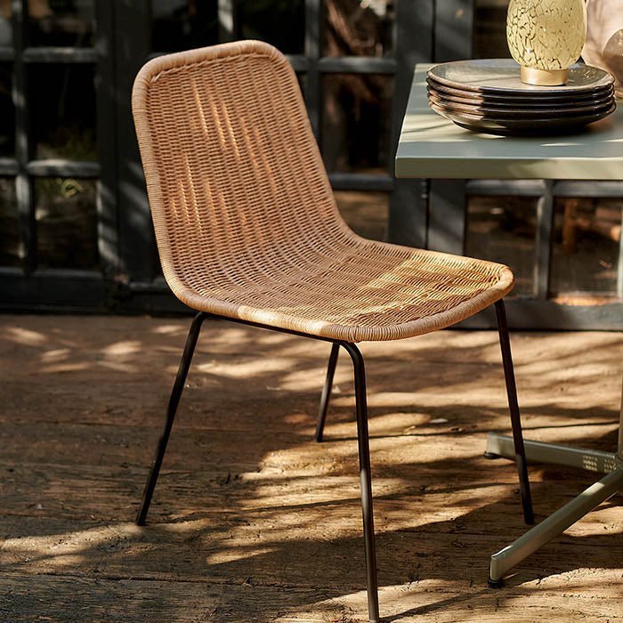 The Tatum Chair, a natural rattan woven chair in a contemporary style with slim black metal legs, on a sun dappled patio.