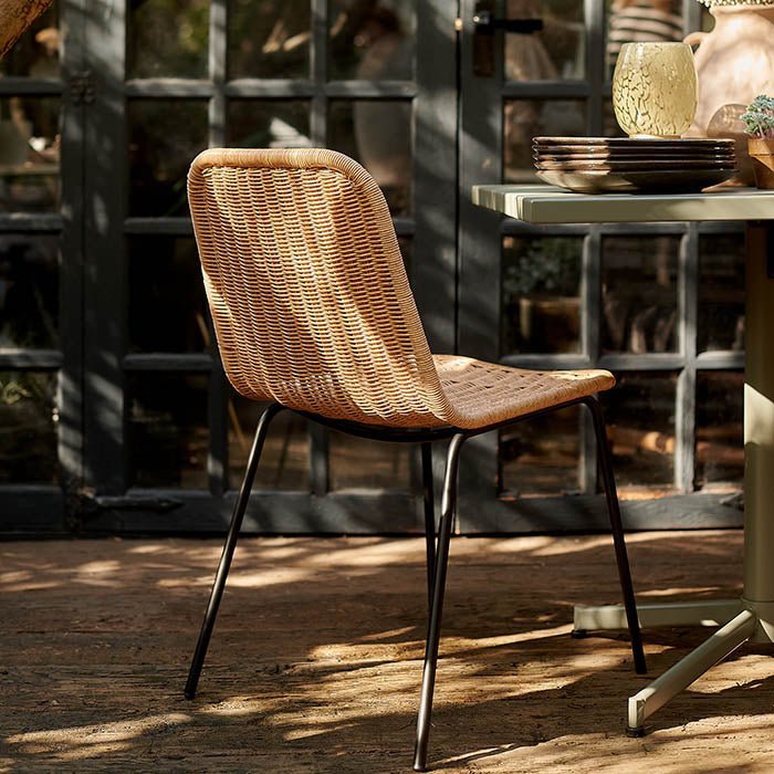 The Tatum Chair, a natural rattan woven chair in a contemporary style with slim black metal legs, on a sun dappled patio.
