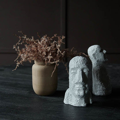 Styled image of two white abstract figurative sculptures next to a small brown ceramic vase with some fake flowers.