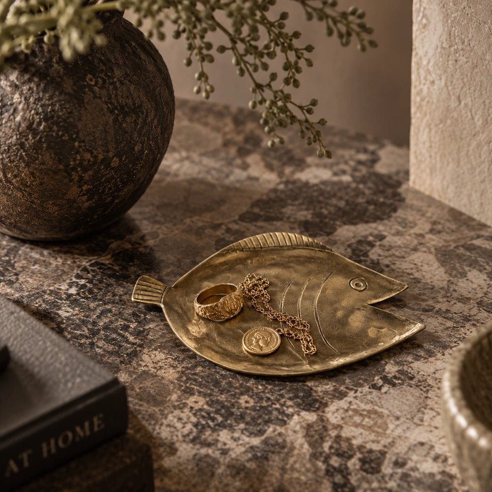 Torcino Brass Tray with organic irregular outline and aged golden finish, displayed on a textured surface beside books, a rounded vase and dried foliage.