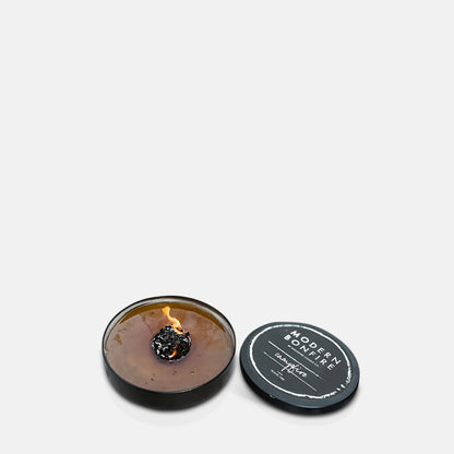 A burning round scented candle in a black metal tin with its lid to the side