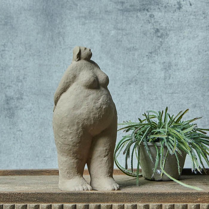 Tall grey standing female sculpture with arms behind her back next to a trailing artificial cactus