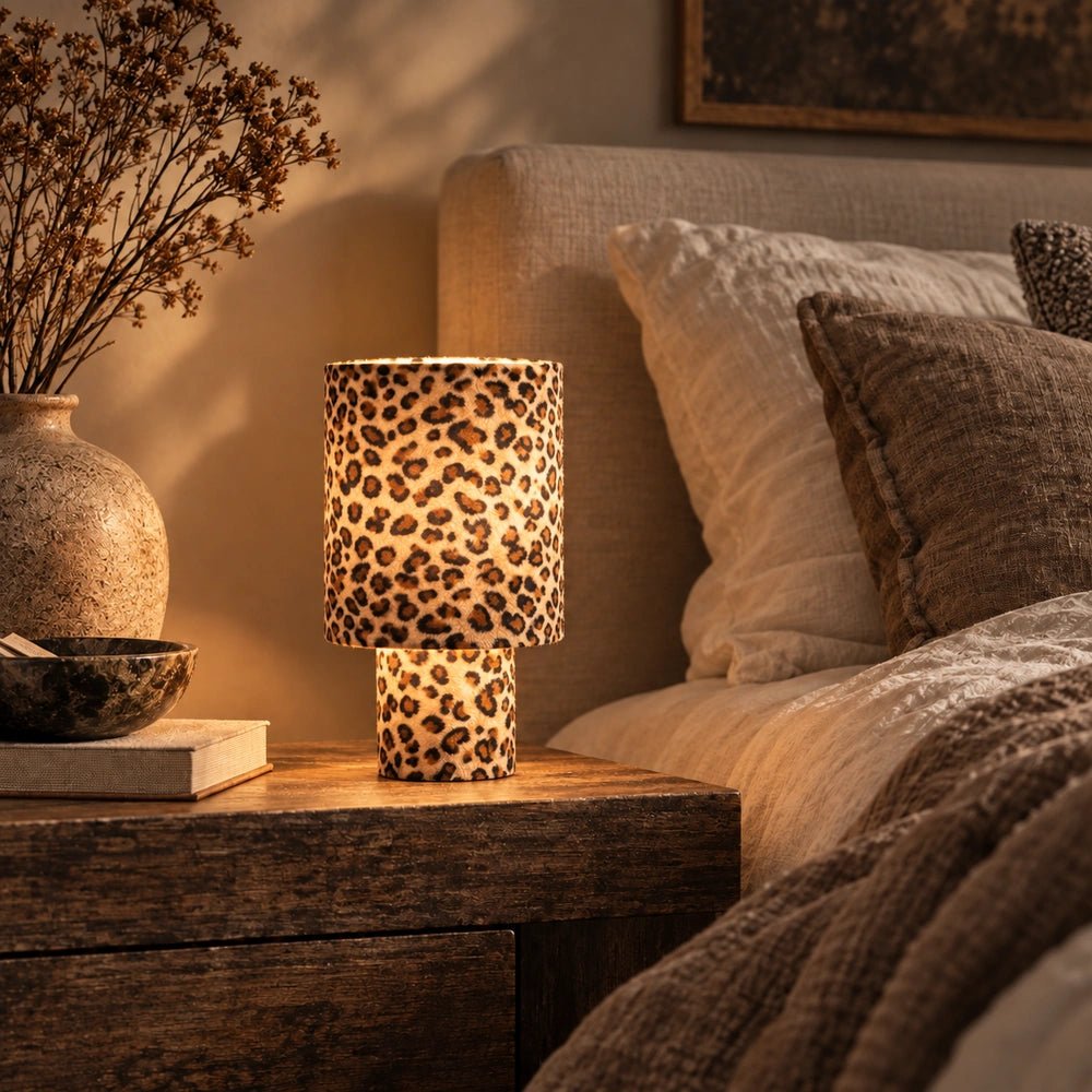 Velma Leopard Table Lamp with spotted cylindrical shade glowing beside a neutral bed, paired with a textured ceramic vase and softly layered brown bedding.