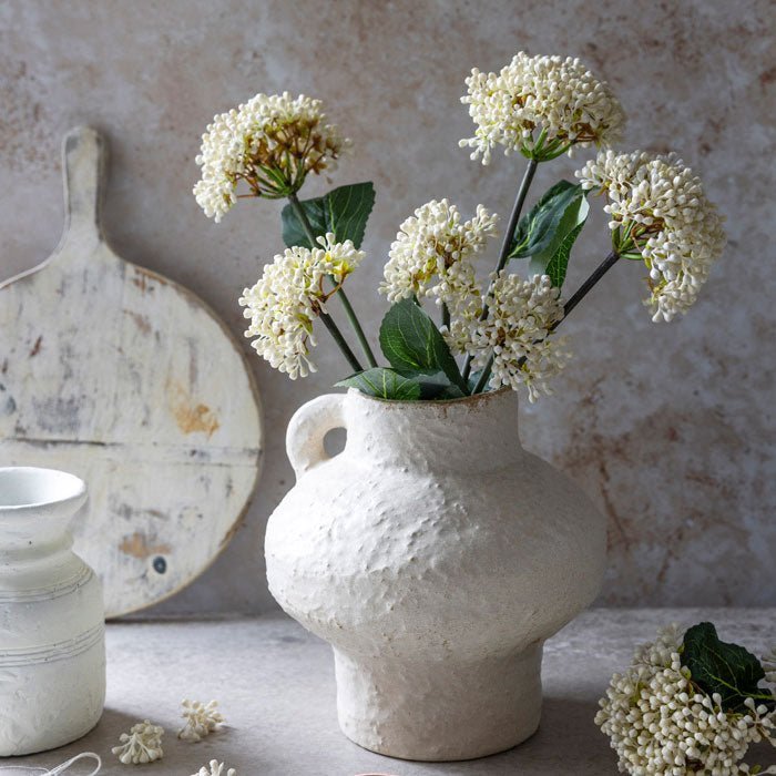 A floral arrangement of cream floral bud stems in a cream ceramic vase.