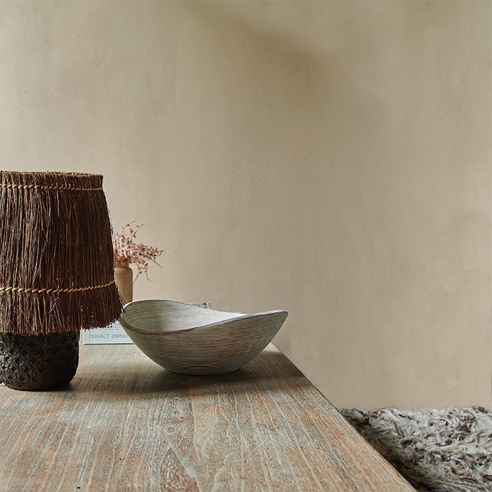 Long wooden table with a brown table lamp and white moon shape bowl against a sandy brown painted wall. Earthy paint colours from Abigail Ahern.