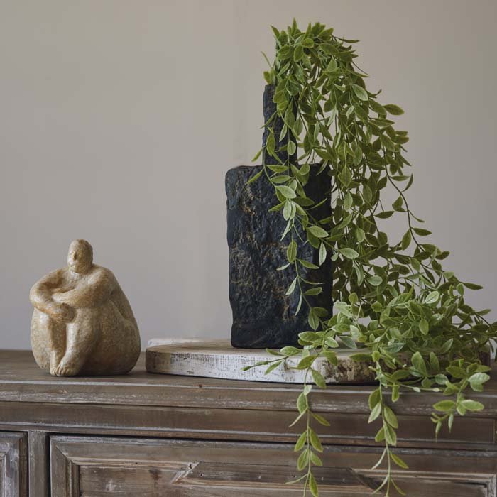 A small grey ceramic vase holding strands of green trailing artificial foliage, next to a small ceramic figurine.