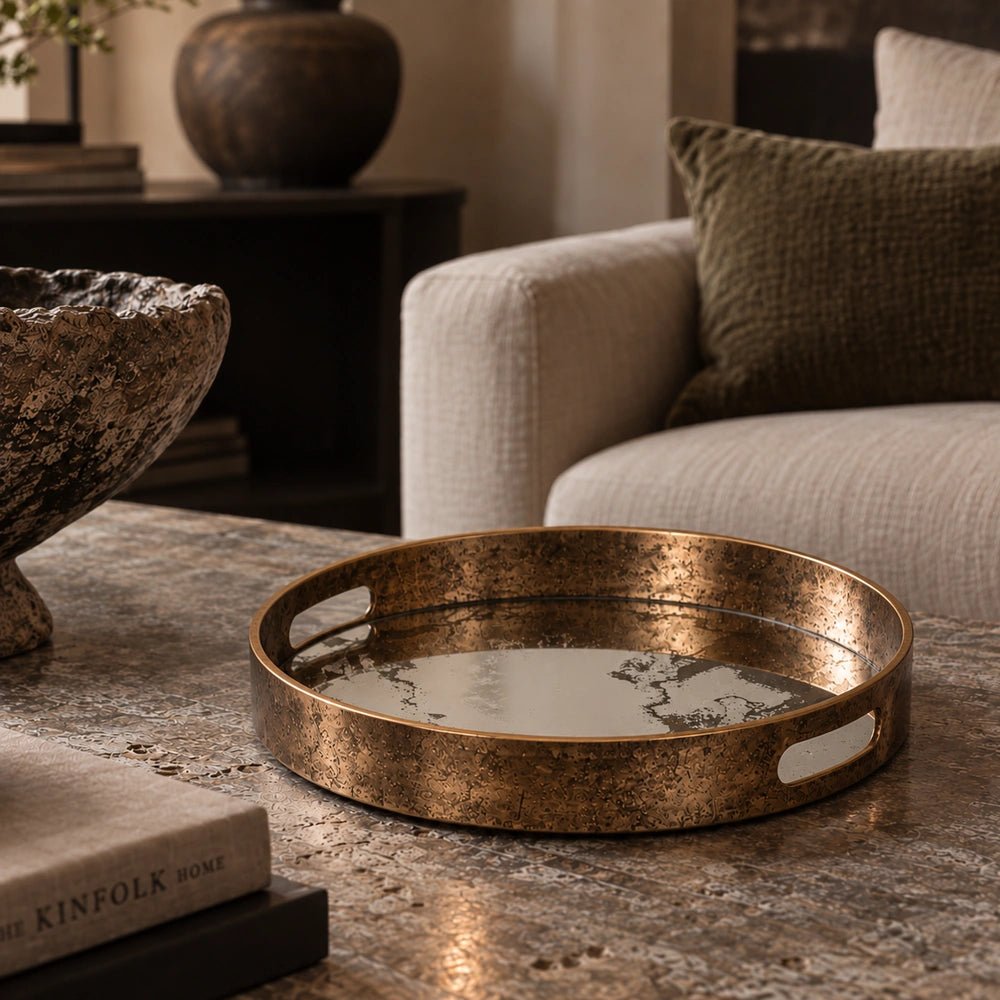 Wilton Tray with round aged brass finish and raised rim, styled on a stone coffee table beside neutral seating, books and dark decorative accessories.