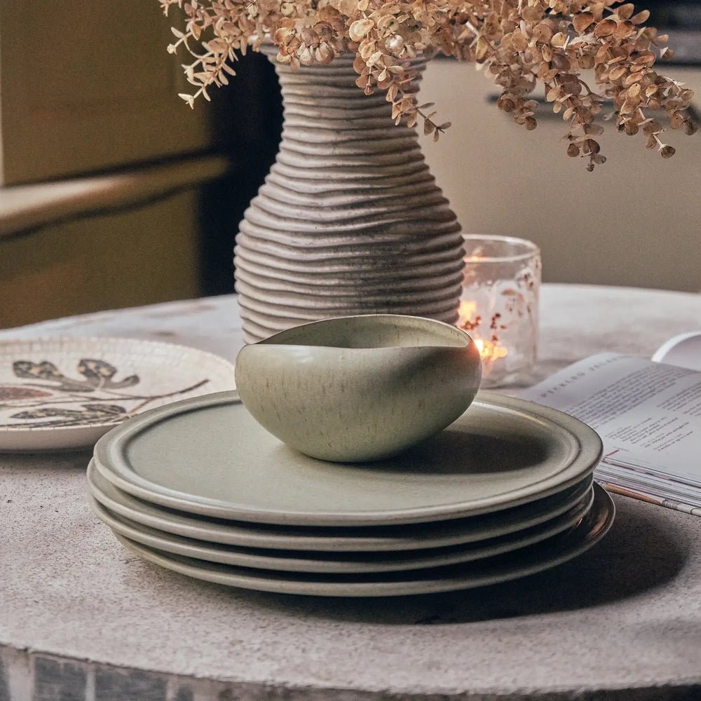 Alcott 12-Piece Dinner Set shown close up with a green rounded bowl above stacked plates, revealing speckled surfaces and raised rims beside a botanical vase.