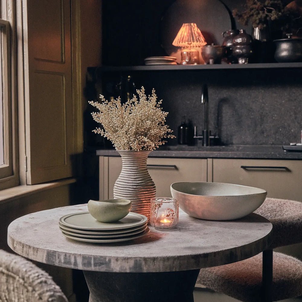 Alcott Serving Bowl with a wide curved grey form, displayed on a round kitchen table alongside stacked plates, a small green bowl and glowing candle.