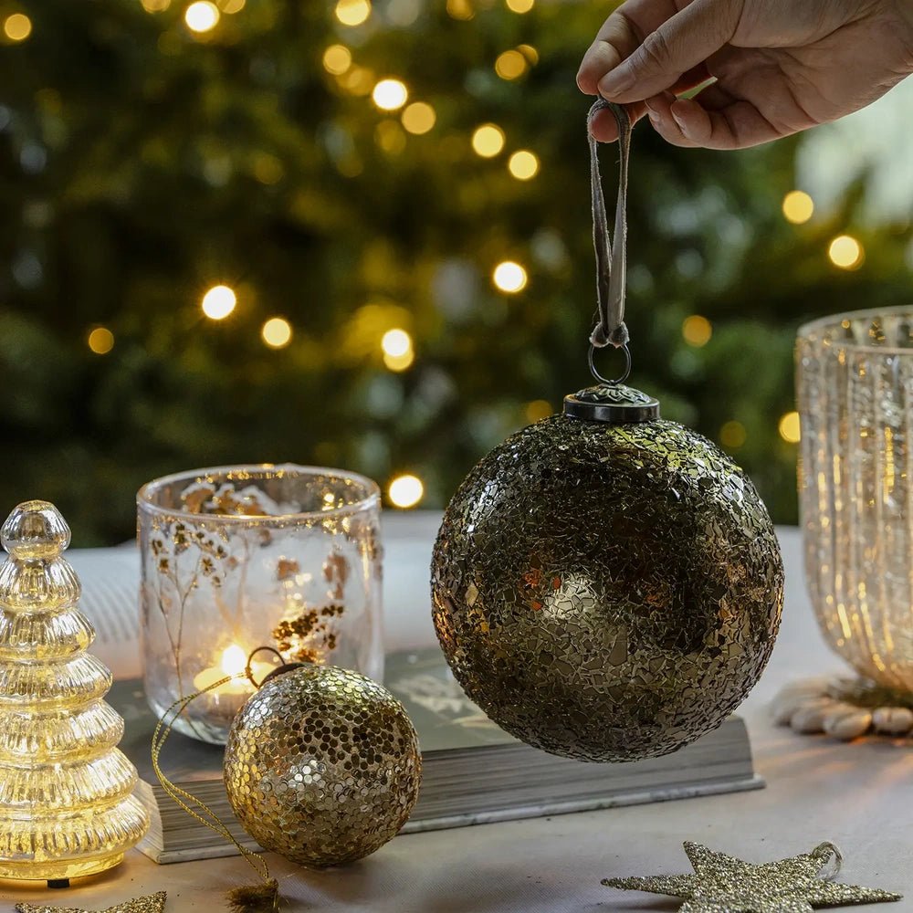 Alisa gold bauble hangs from a loop held above a festive table, displaying its round textured surface beside smaller ornaments and a candleholder.