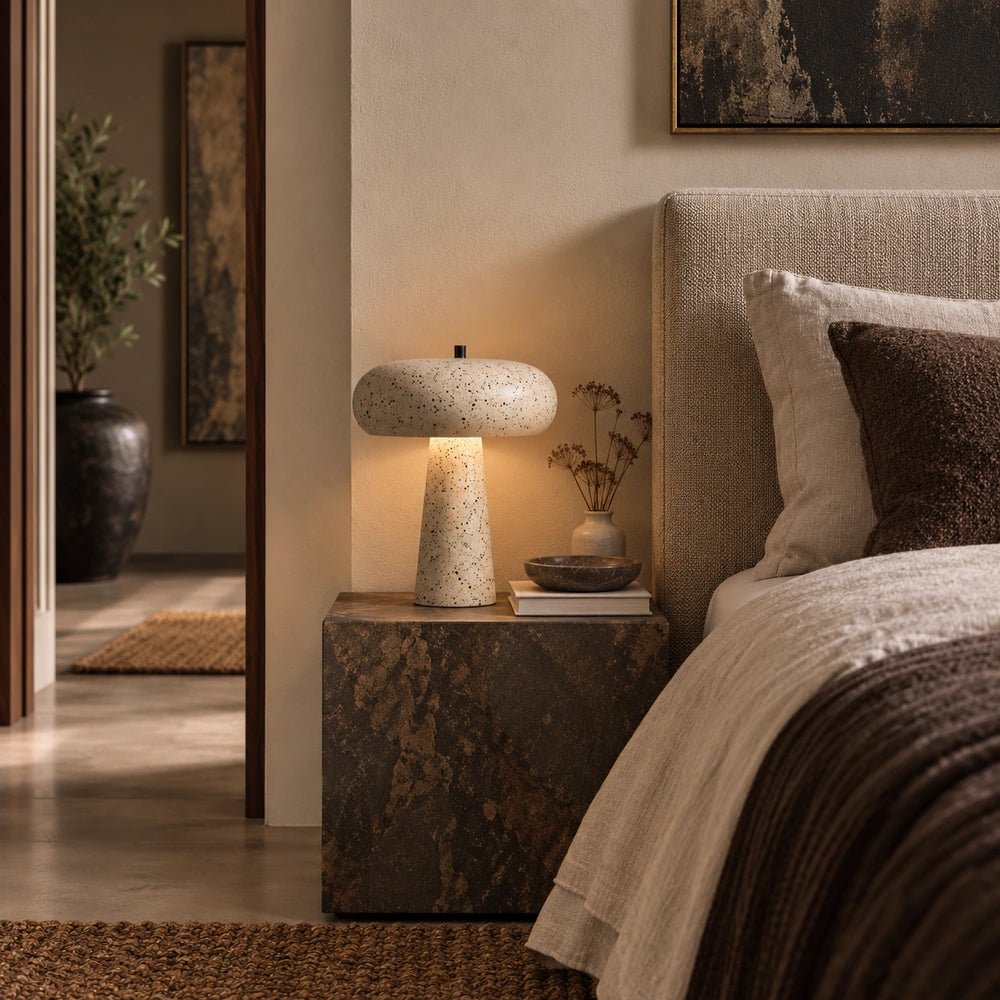 Alma Metal Table Lamp with speckled rounded shade and tapered base glowing on a dark bedside table beside layered neutral bedding.