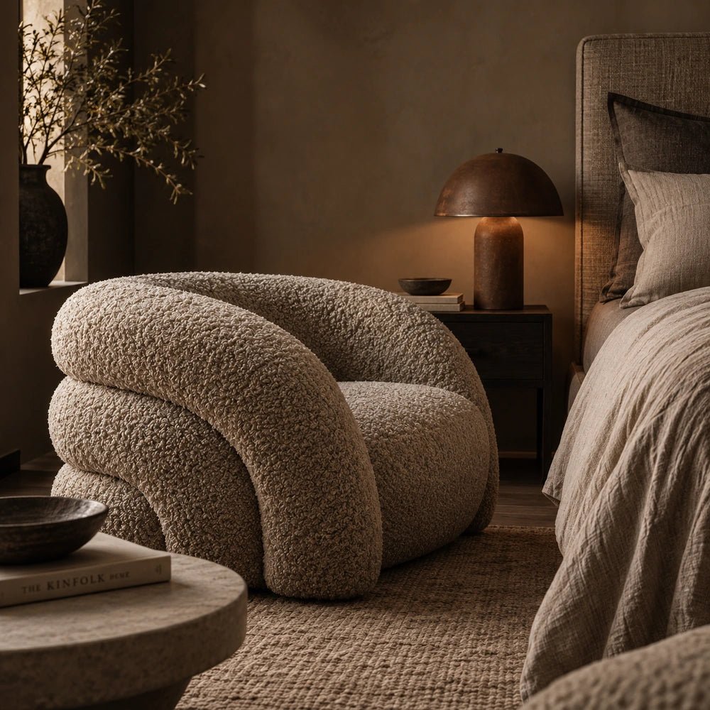 Almar Curved Armchair with rounded textured upholstery beside a bed, framed by warm neutral walls, dark bedside furniture, layered bedding and soft ambient lighting.