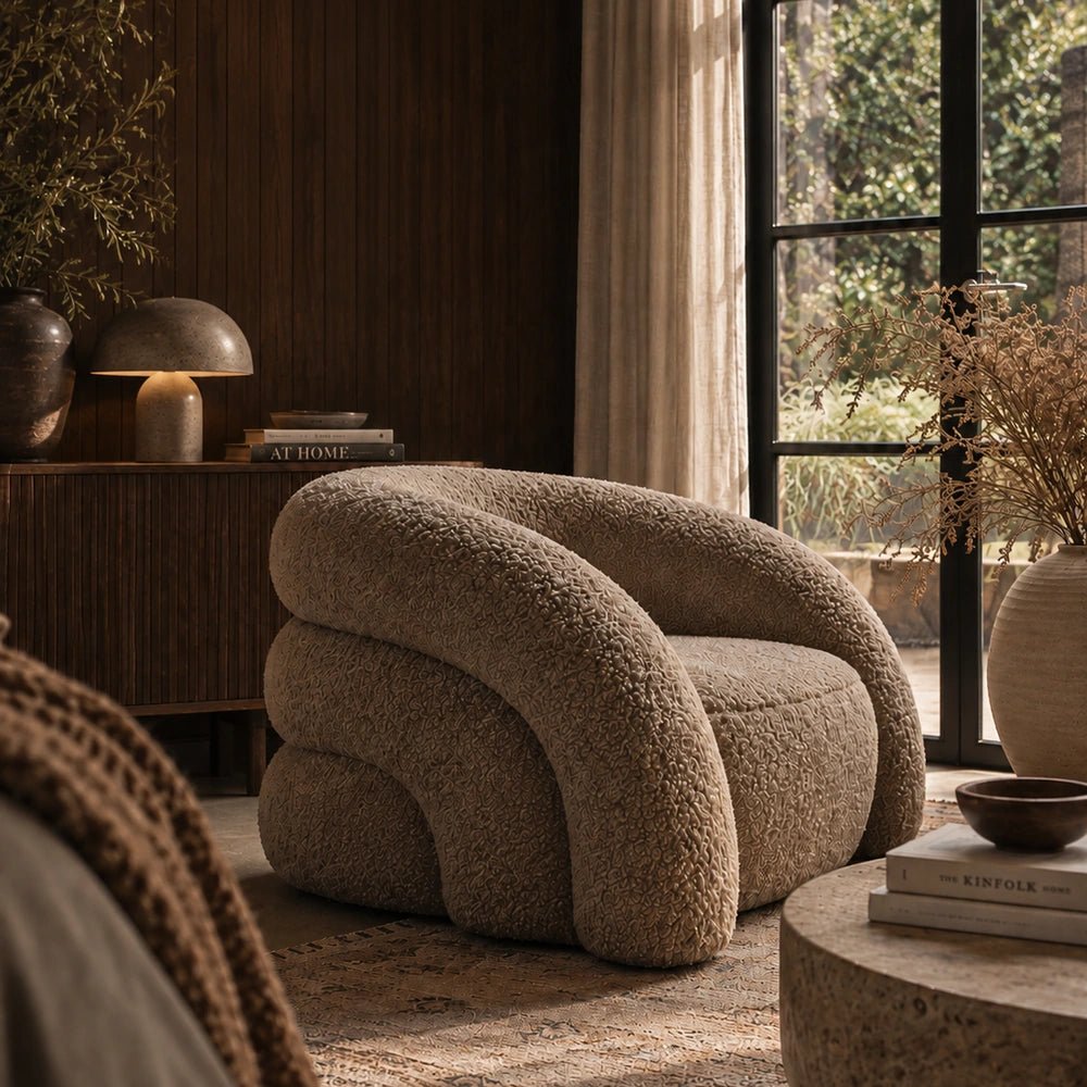 Almar Curved Armchair in textured taupe upholstery, shown at a left angle in a warm living room with dark wood furniture, rug and window light.