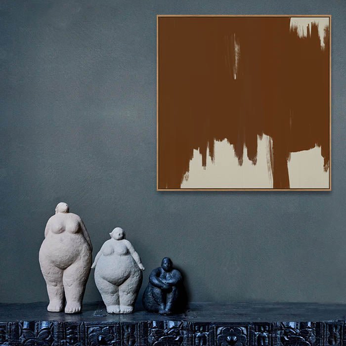 Square brown and cream print in thin gold frame hung above three female sculptures