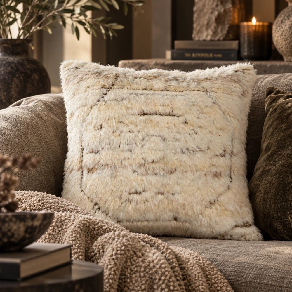 Astoria Wool Cushion with soft shaggy neutral surface, styled among layered cushions on a sofa in a warm contemporary living room setting.